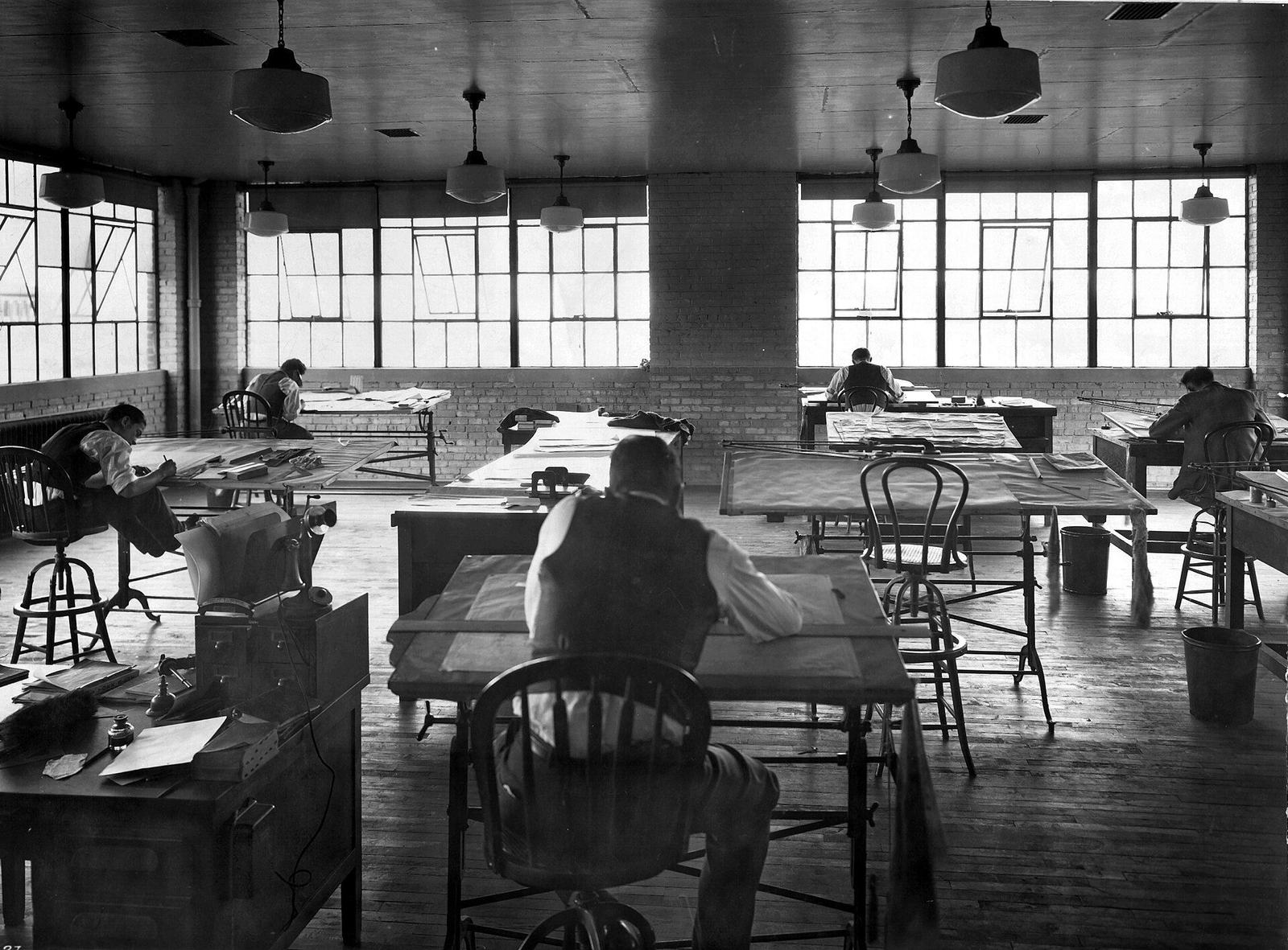 Large drafting room with draughtsmen at tables under hanging lamps