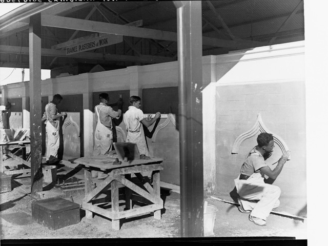 Trainee plasterers in work clothes shaping ornamental plaster on practice walls in a training shed