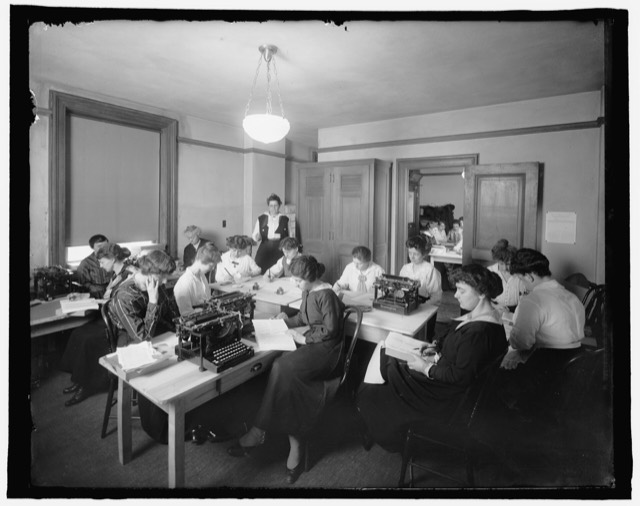 Women working at typewriters around shared tables in an office from the 1910s