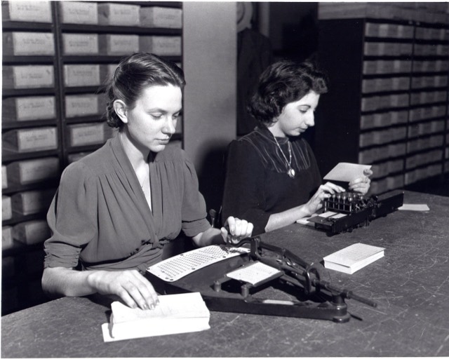 Two clerks at the US Census Bureau punching population cards on a pantograph and a keypunch, with filing boxes behind them