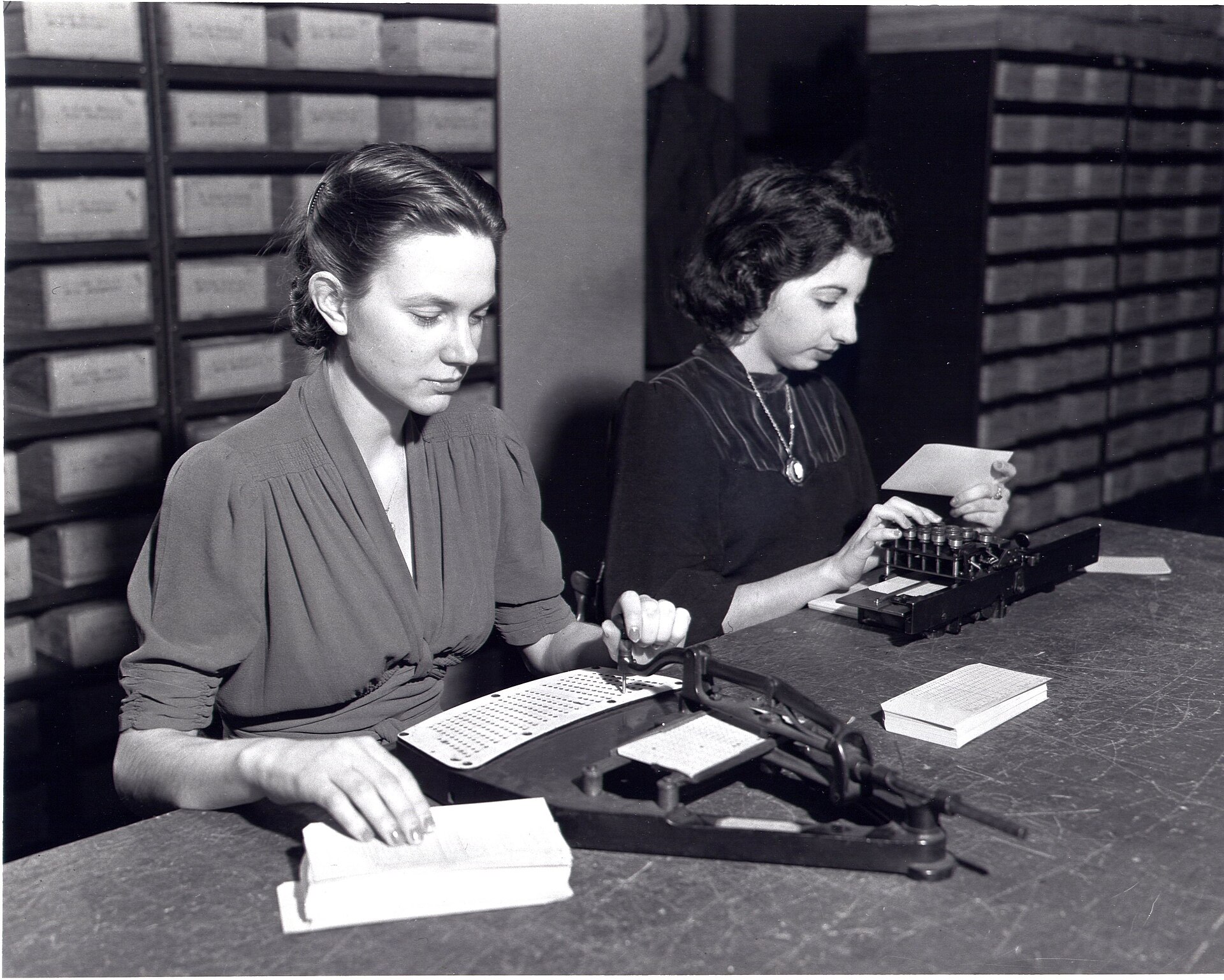 Two clerks at the US Census Bureau punching population cards on a pantograph and a keypunch, with filing boxes behind them